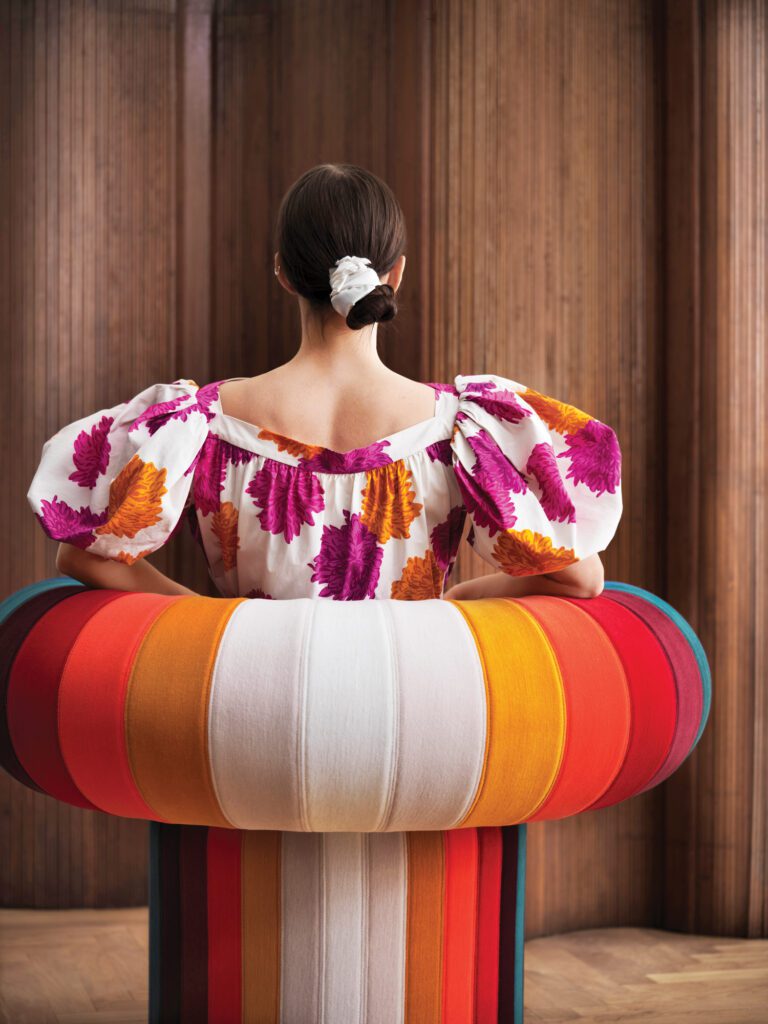 Big Talk, Adam Goodrum Studio’s semicircular throne for Blå Station, pictured here, is a rounded arm chair decorated with vertical stripes in blue, shades of red, gold and white. A woman sits in the chair with her back to the camera. 