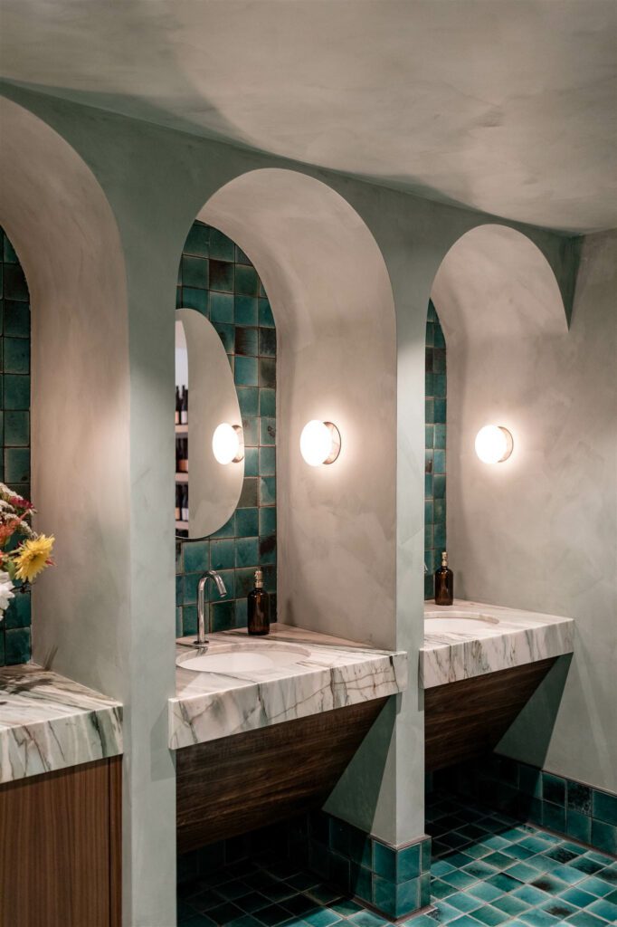 Allied Maker frosted disc and orb sconces shine bright on each side of the personal vanities in a restroom featuring Cle Tile’s Eastern Earthenware collection and Portola Paints lime wash in Rococo.