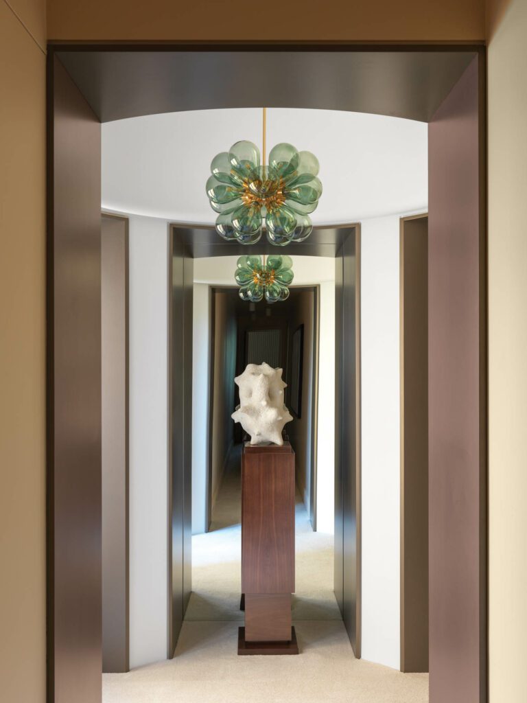 The corridor leading to the primary bedroom hosts a Tom Kirk chandelier and a William Coggin stoneware sculpture, which graces a walnut plinth.