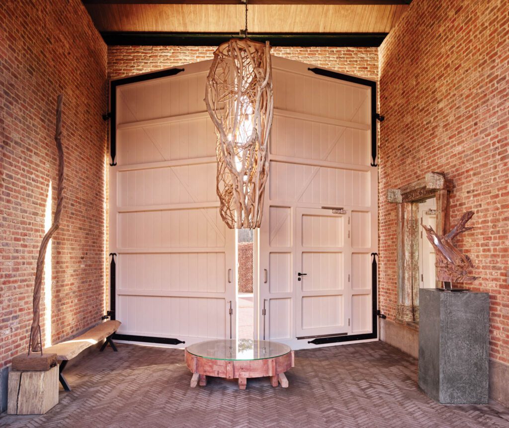 A custom chandelier made of local tree branches and a 19th-century Indonesian door repurposed as a mirror furnish the main entry to a stable and a private residence in Retie, Belgium, renovated by Sceners.