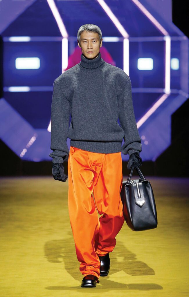 A male Prada model in orange pants and a gray turtleneck. 