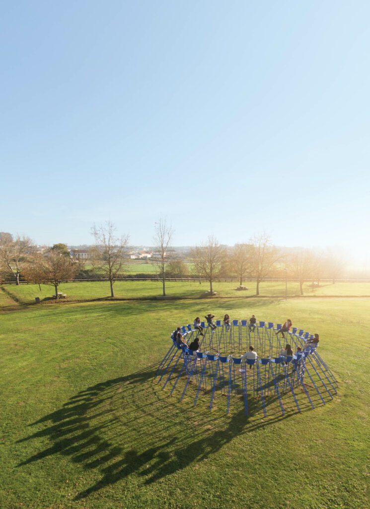 visitors sit in blue steel stacking seats in a circle in the middle of an open field