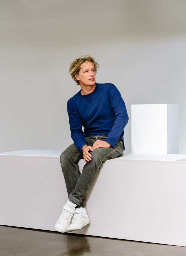 Yves Béhar in a blue long-sleeve shirt sitting on a white ledge. 