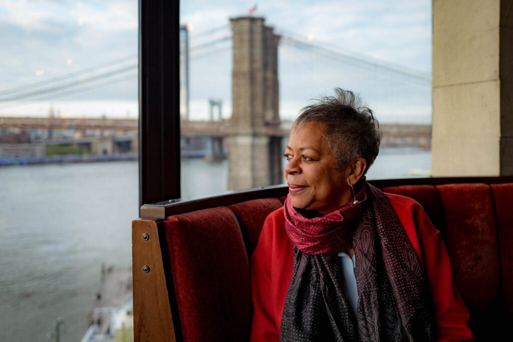 Mavis Wiggins wearing a red scarf looks out at the waterfront neat the Brooklyn Bridge.