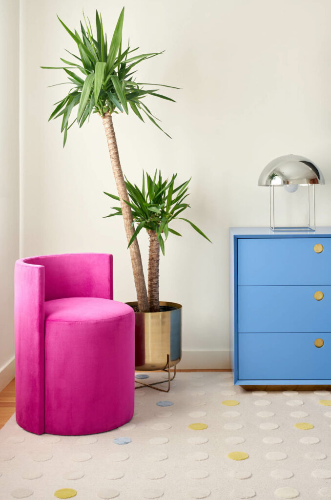 A bright pink armchair pops next to a palm tree on a rug with subtle yellow and cream circles.