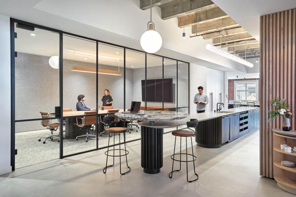 An office kitchen with an island and a high top table with two stools near a conference room. 