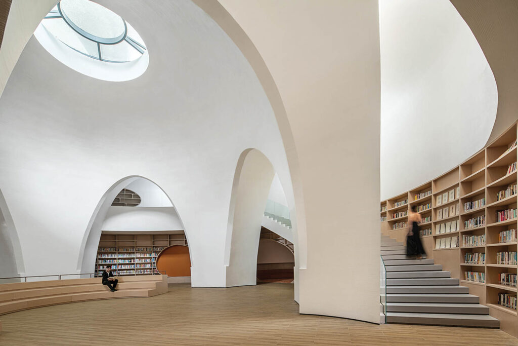 A man sits on curved benches in front of shelves of books and a woman in a black dress walks up a staircase beneath sweeping white arches.