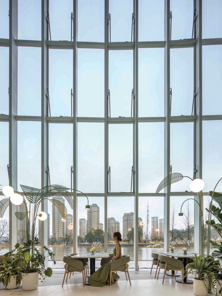 a woman sits at a dining table in front of a glass wall surrounded by greenery