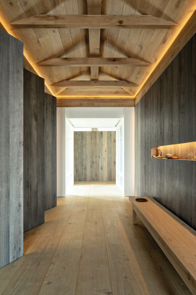 wide-plank French-oak flooring stained warm gray and cove lighting turns a passageway’s beamed ceiling into a floating, poetic vision