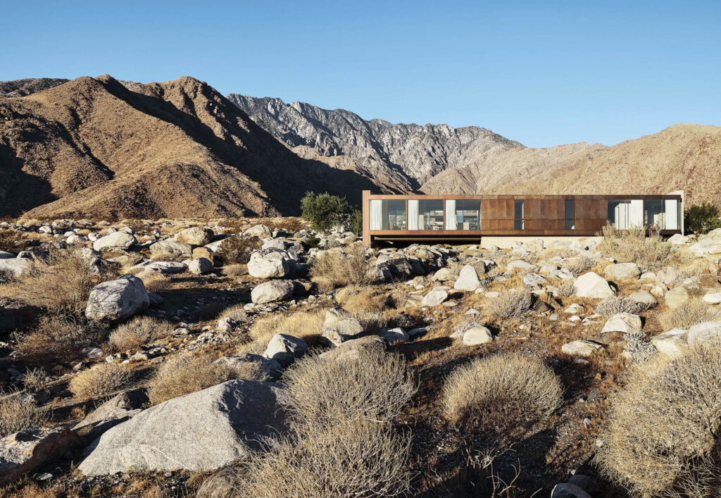 A view of the home's rectangular wooden and glass exterior, which blends in with the mountains behind it and the desert landscape. 