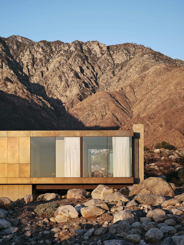 A view of the home perched on surrounding boulders with floor-to-ceiling glass windows covered by cream curtains. 