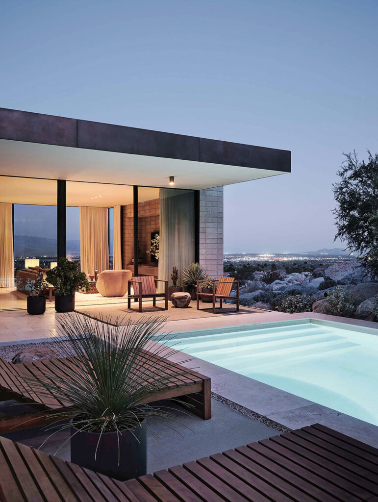 The living area opens to the pool deck at dusk with a wooden lounge chairs.