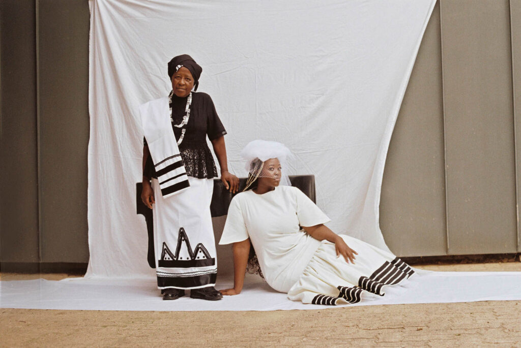 Thabisa Mjo sits on the floor in front of a table while her mother stands nearby. 