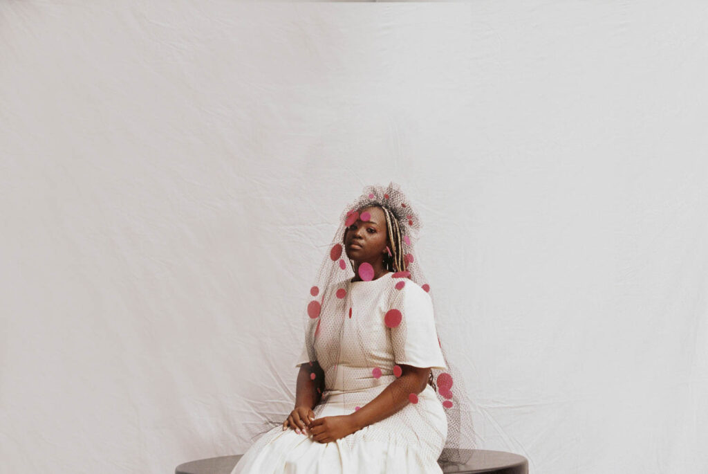 Thabisa Mjo wearing a sheer veil with red dots throughout and a white dress sitting on a table. 