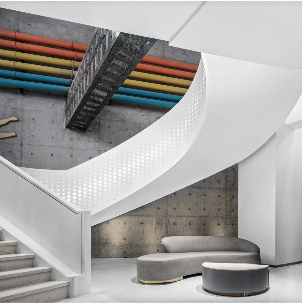 A white curving staircase with rainbow hued industrial pipes above.