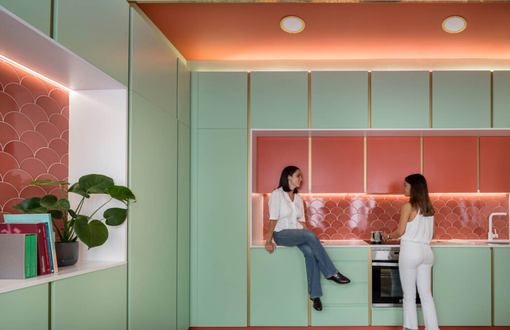 A woman sits on a light green countertop talking to a friend in an apartment