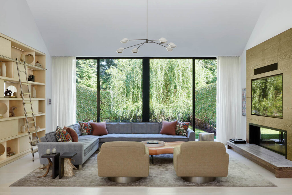 a living area with a sectional and accent chairs with a glass wall with views of trees