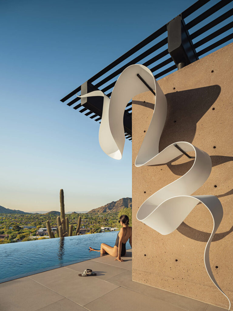 woman sitting by edge of pool underneath a brown building with white sculpture