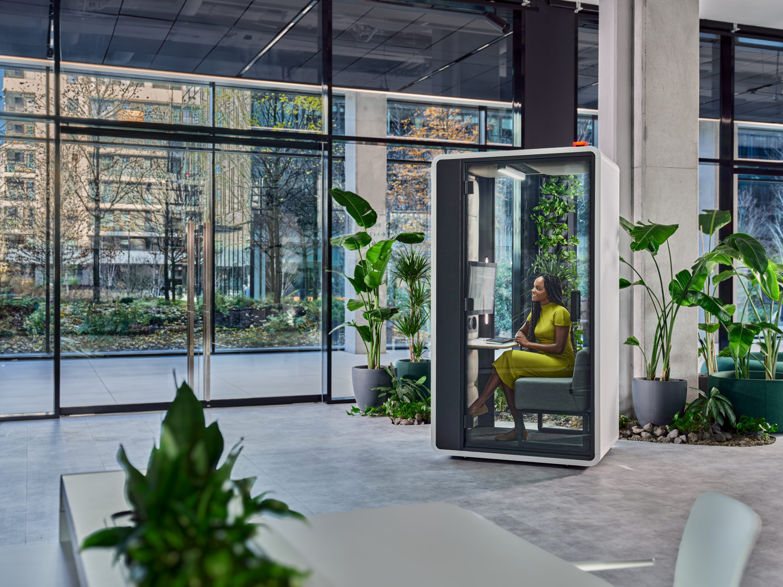 Woman seated inside a modern acoustic office pod in a bright, plant-filled workspace with floor-to-ceiling windows.