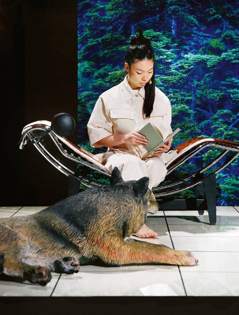 A woman sitting on a chair reading a book next to a bear.