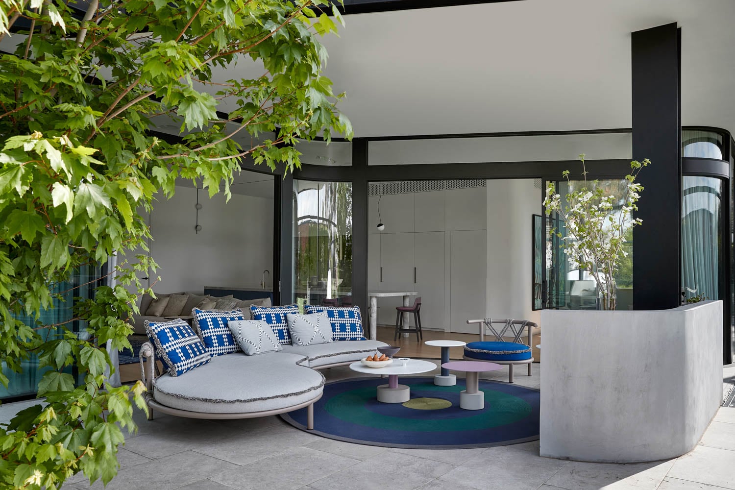 Modern outdoor lounge with round couch, blue-patterned pillows, circular tables, and lush greenery