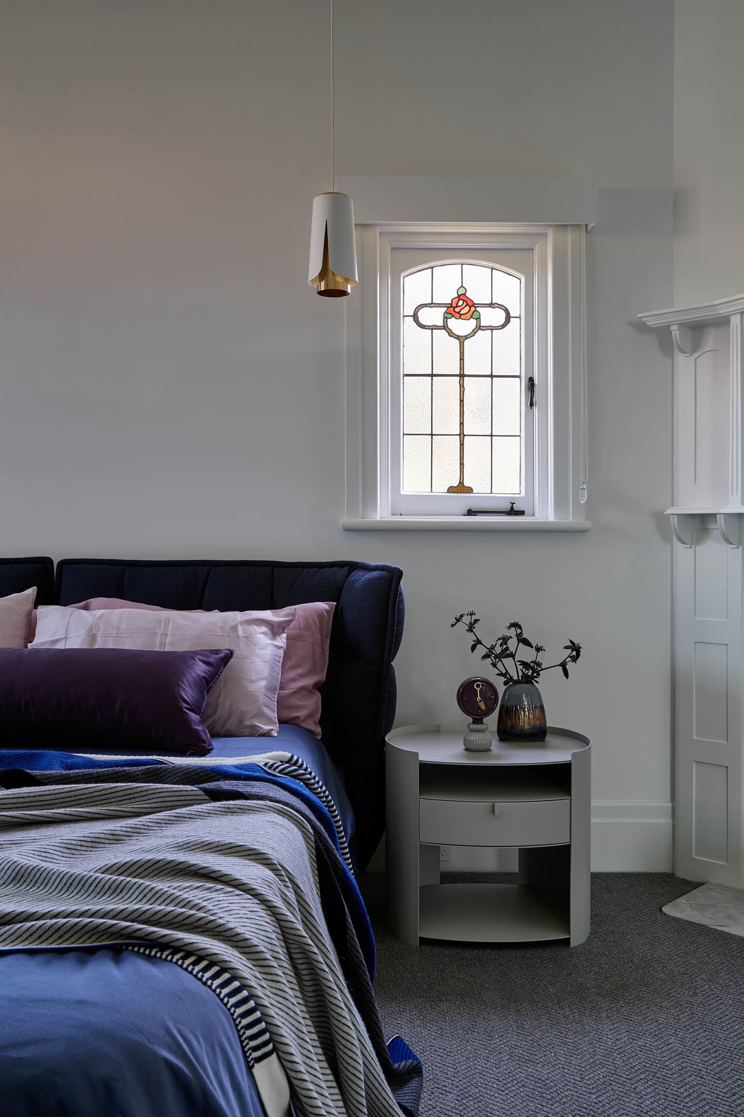 Cozy bedroom with a deep blue bedspread, purple and pink pillows, and stained glass window