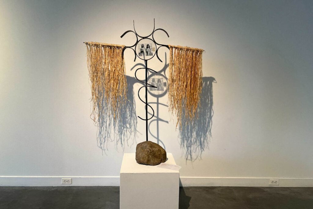 A metal sculpture on a white pedestal features curving shapes, straw-like tassels, and small images.
