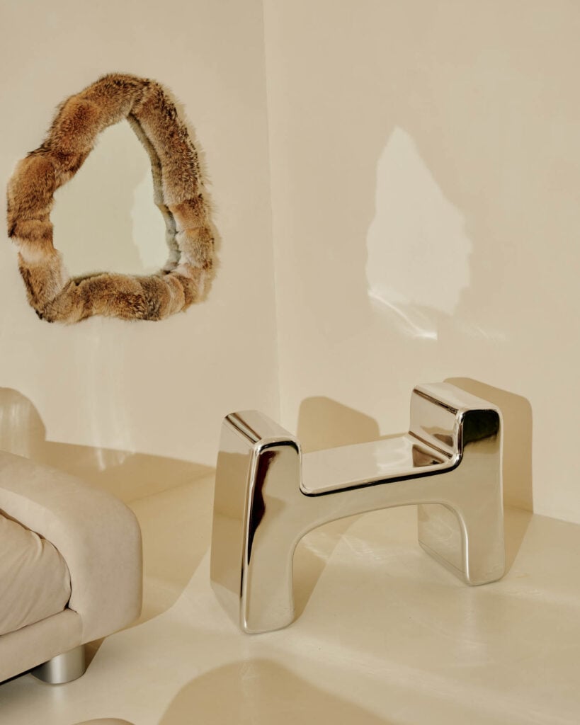 Futuristic chrome stool beside a beige sofa and fur-framed mirror. Minimalist interior, soft lighting, modern elegance, warm tones.