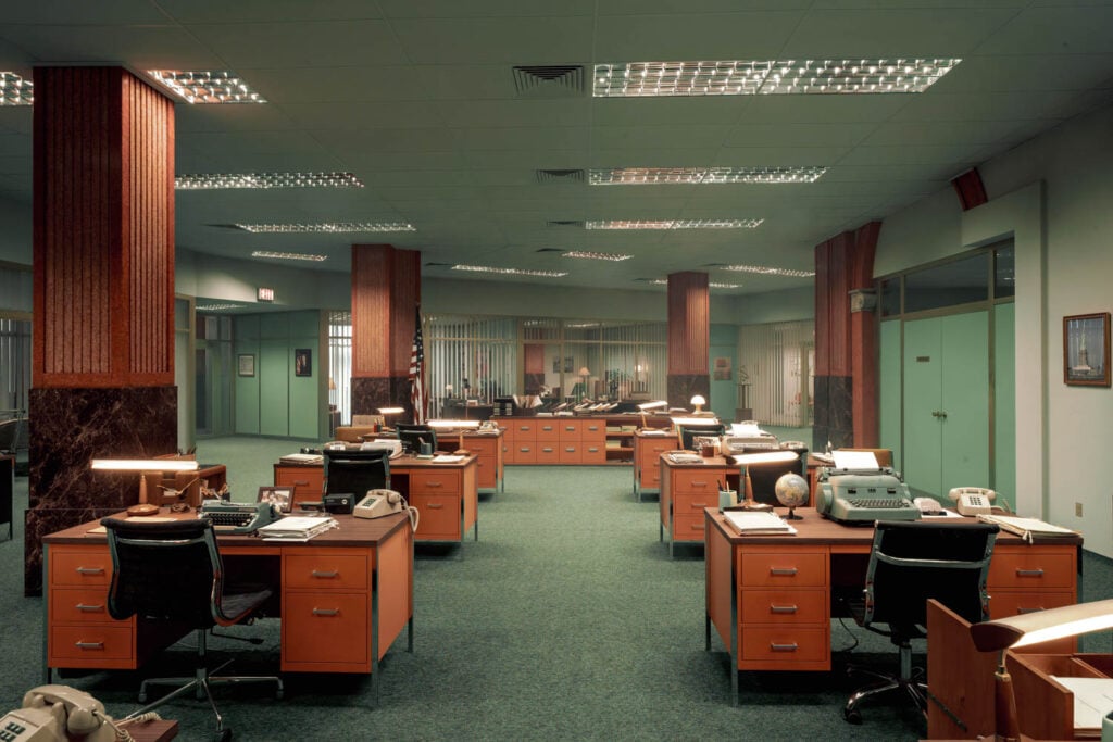 Spacious vintage office with rows of wooden desks and rotary phones, dimly lit by fluorescent lights