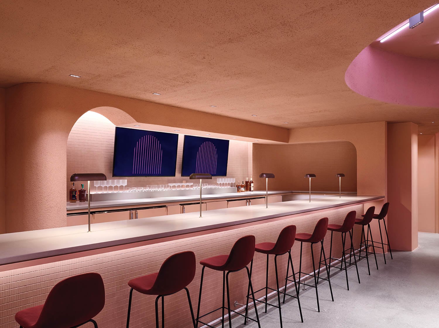 Modern bar interior with pink hues, featuring sleek bar stools and minimalist lamps along a long bar counter