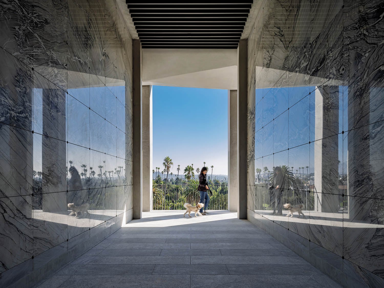 A person walks a dog through a marble hallway toward an open view of palm trees under a blue sky