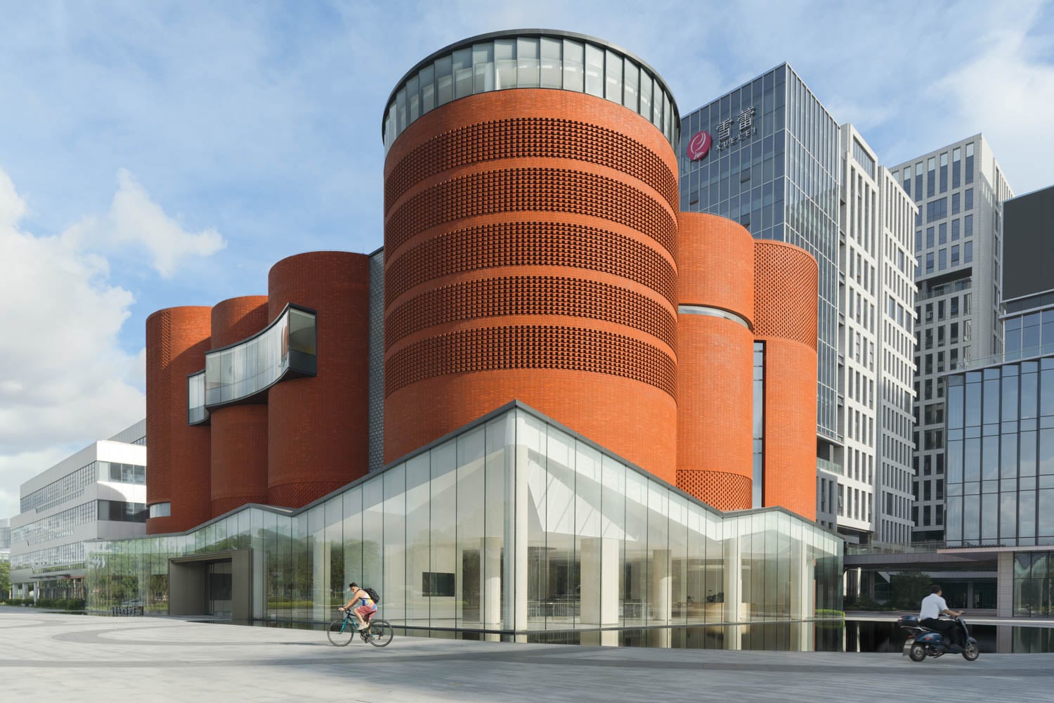 Modern building with cylindrical red brick structures and glass façade, set against a clear sky