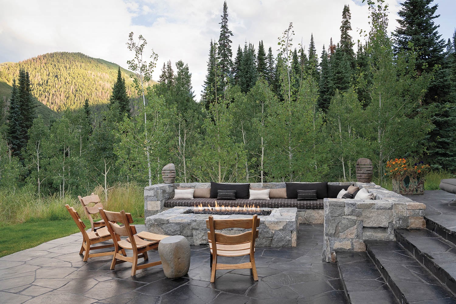 Outdoor patio with stone fire pit surrounded by wooden chairs and cushioned seating