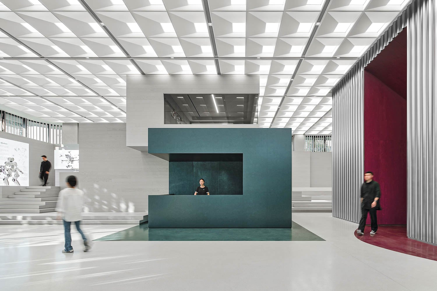 Modern interior with geometric ceiling, a central dark turquoise reception desk, and people walking