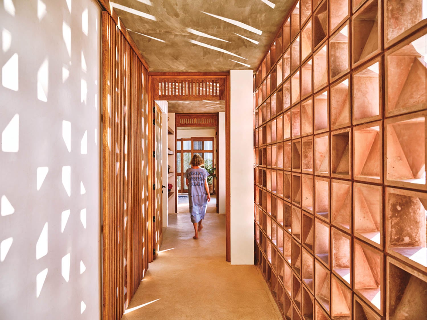 A person walks down a sunlit hallway with wooden lattice walls and geometric shadows