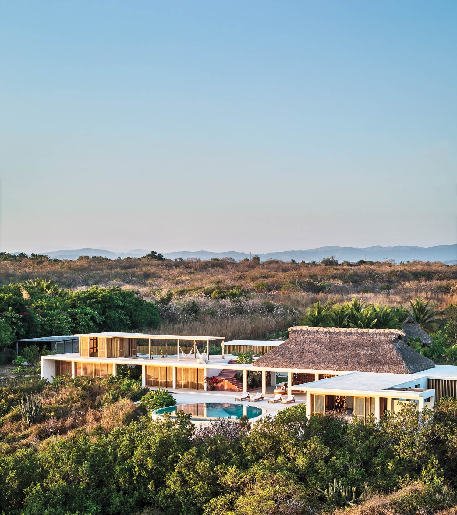Modern villa with a thatched roof and glass walls nestled in lush greenery, featuring an infinity pool