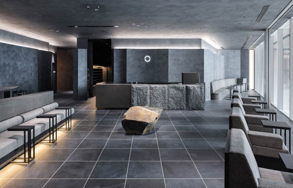 Modern minimalist lobby with gray tones, featuring sleek benches, a textured stone counter, and a large rock centerpiece on a tiled floor