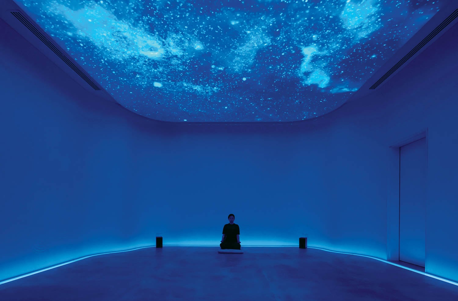 A person sits meditatively under a starry sky projection in a dimly lit, blue room