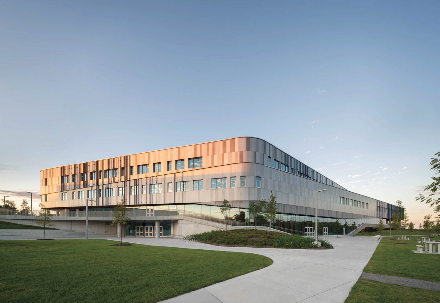 Modern three-story building with metallic facade, large windows, surrounded by green lawns and paths under a clear blue sky at sunset