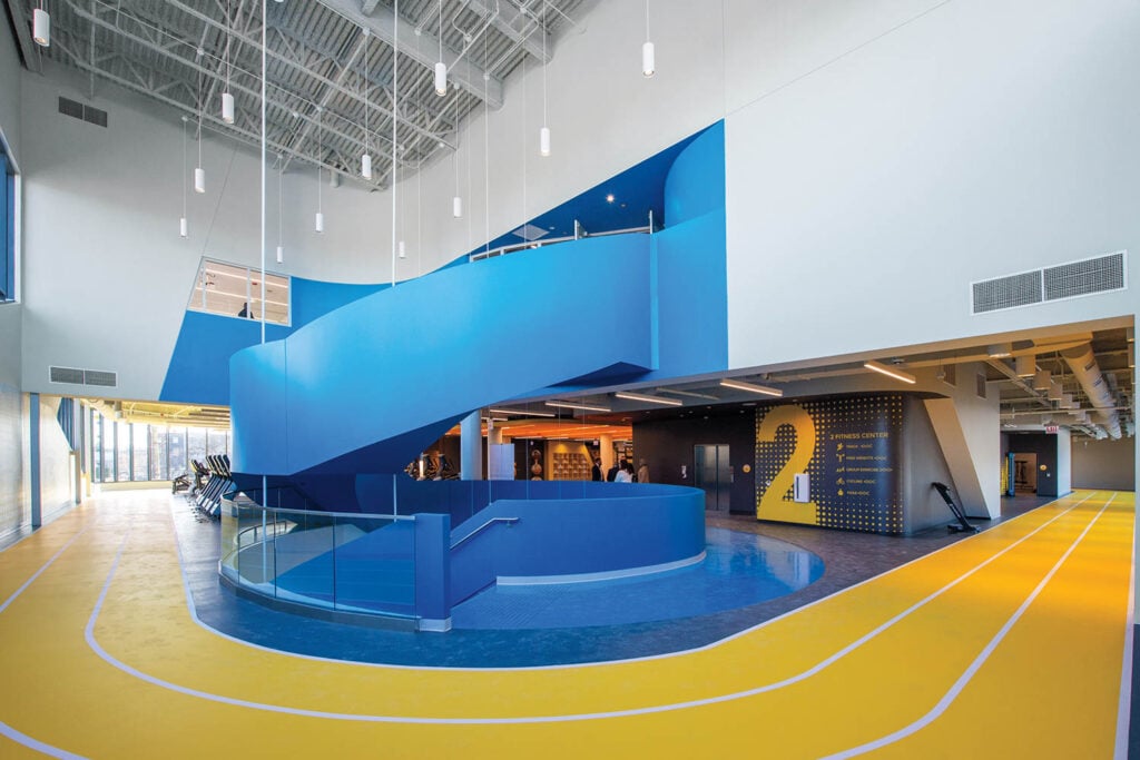 Spacious modern gym interior with a striking blue spiral staircase, yellow running track, and high ceilings.