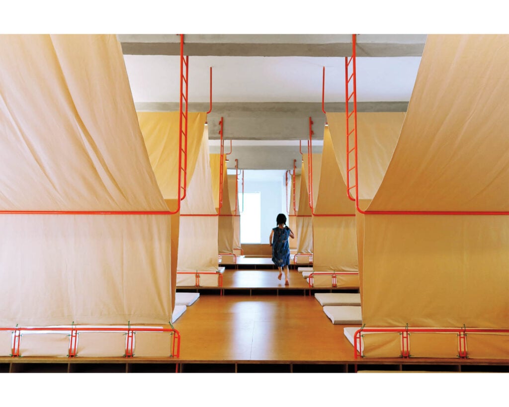 A child in a blue dress walks between a row of hanging orange sleeping pods with red frames in a bright, minimalistic room