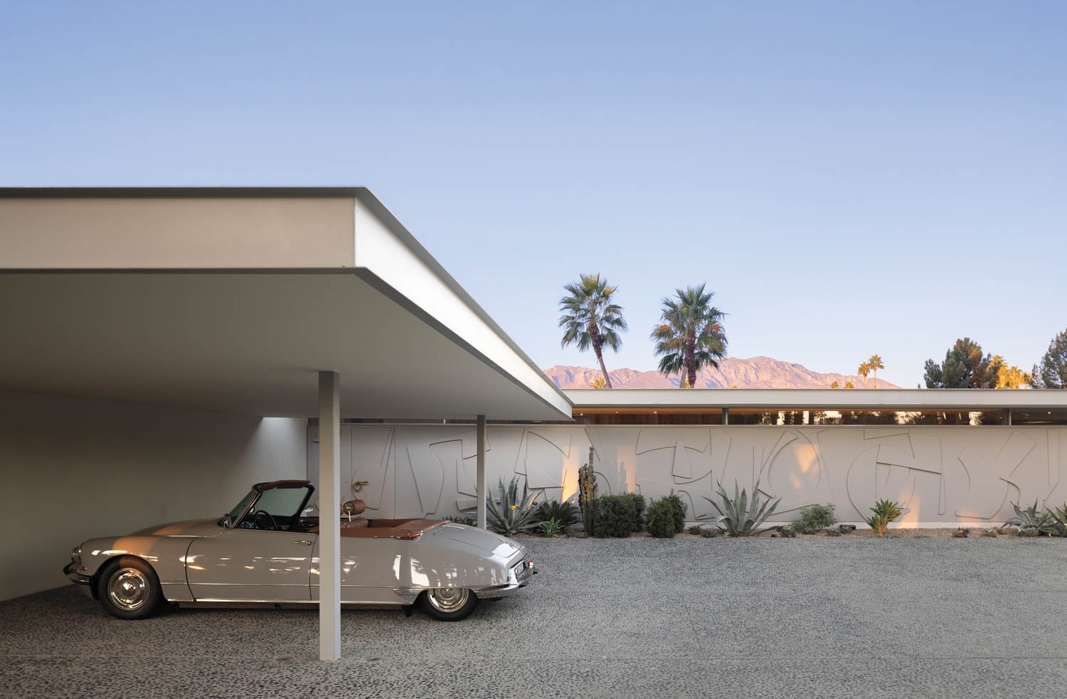 A classic convertible car parked under a modernist carport beside a minimalist house, with palm trees and mountains in the serene background