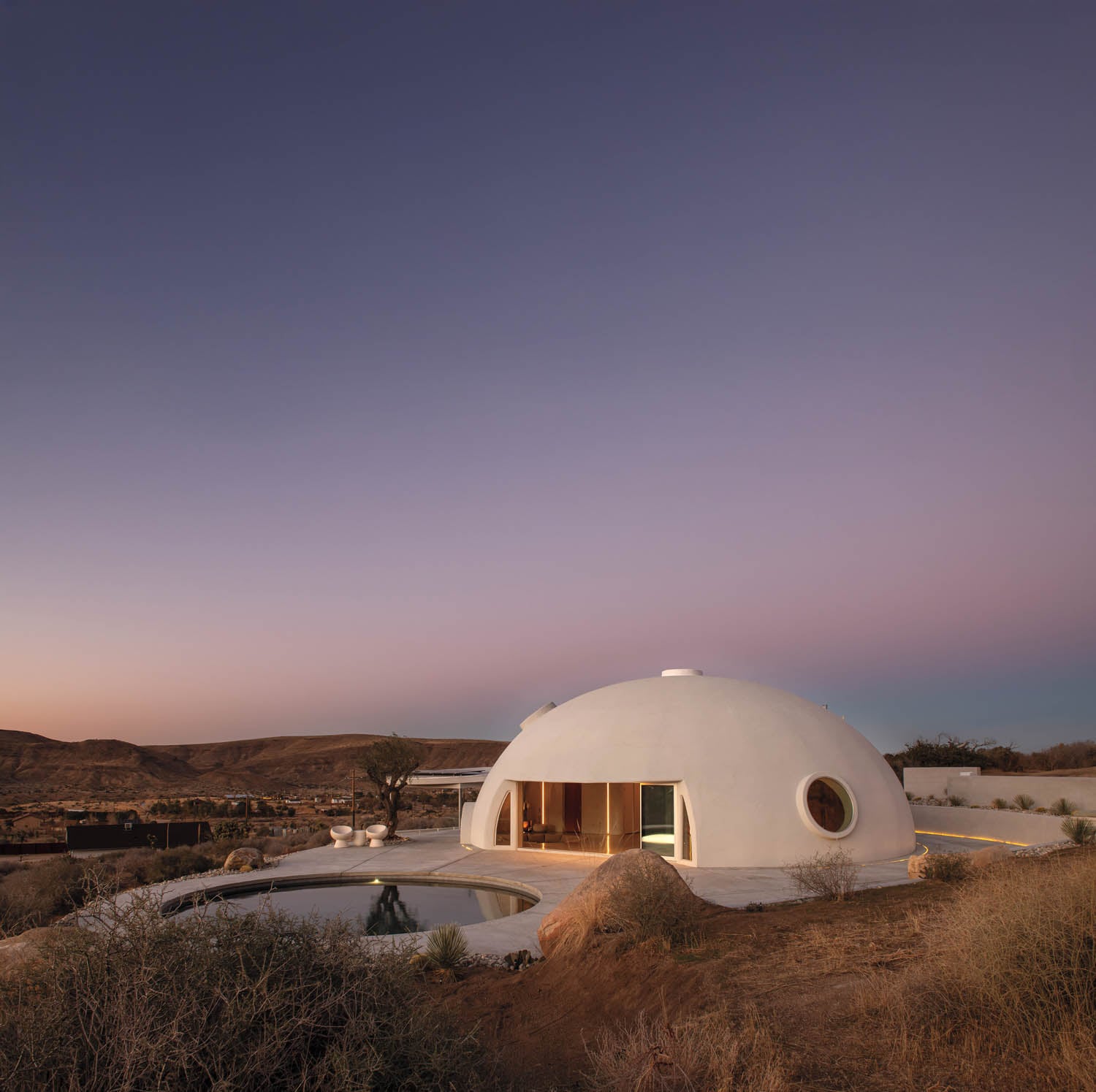 A white, dome-shaped house with circular windows sits in a desert landscape at sunset, surrounded by dry bushes and hills