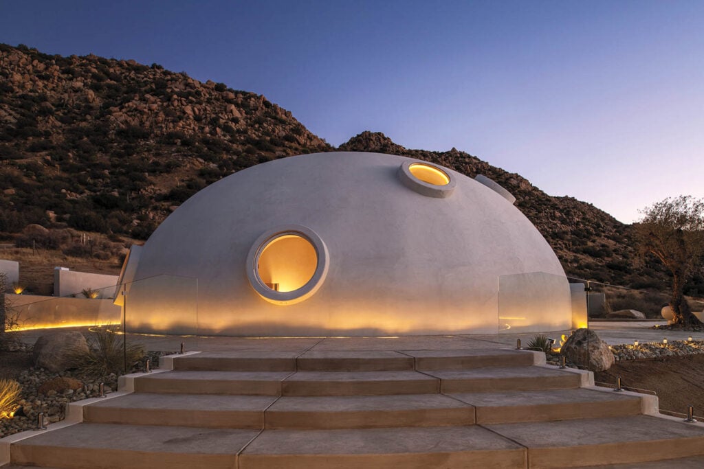 A dome-shaped building with circular windows glows warmly under a twilight sky, surrounded by rocky hills
