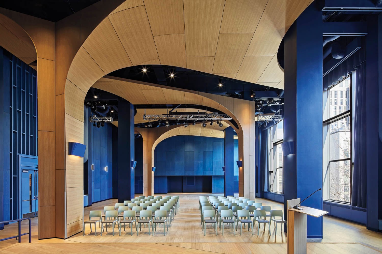 Spacious modern auditorium with curved wooden ceiling, blue walls, large windows, and rows of empty chairs