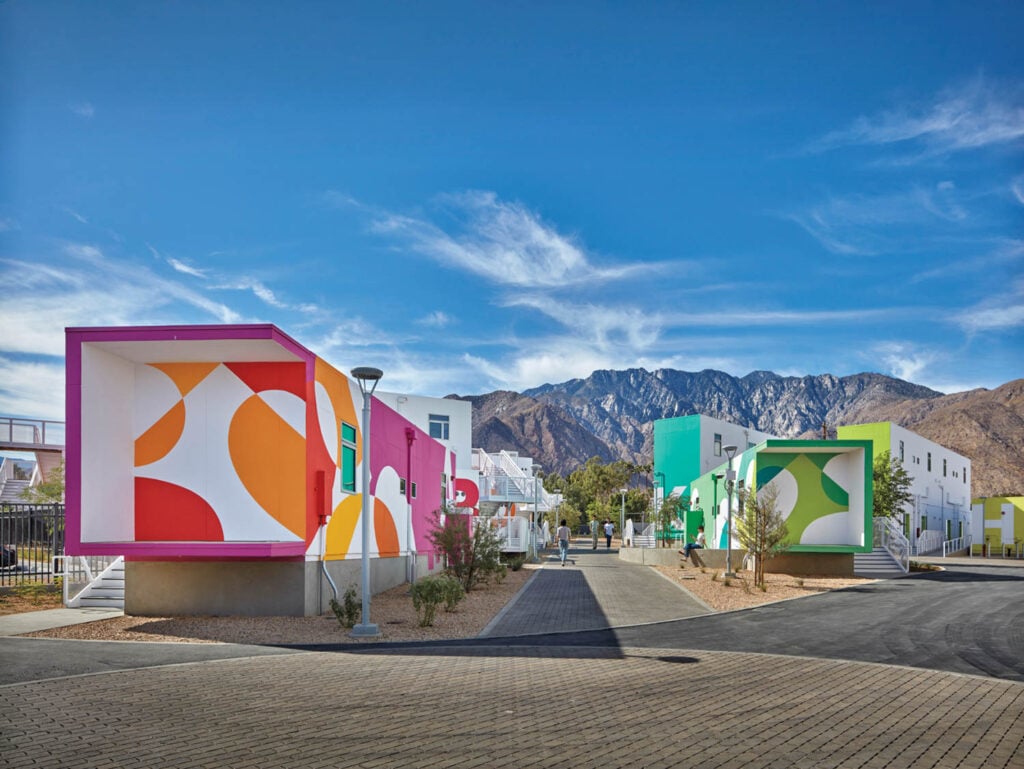 Colorful, modern buildings with geometric patterns line a paved path under a bright blue sky