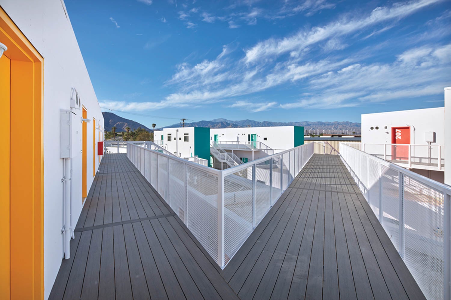 Modern urban rooftop deck with white railings and colored doors, overlooking mountains under a vibrant blue sky