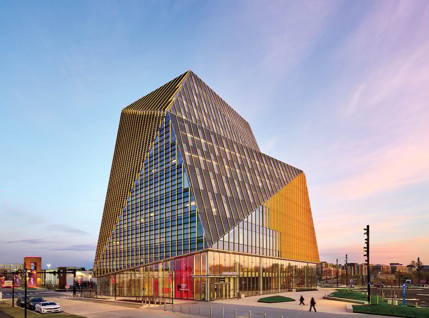 A modern, angular glass building with crisscrossing metal beams stands against a sunset sky