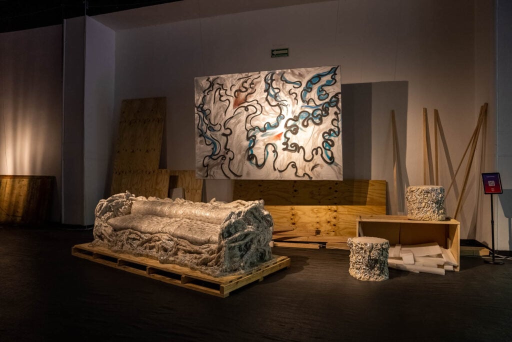 An abstract art installation in a dimly lit room featuring a large painting with swirling black, blue, and orange lines