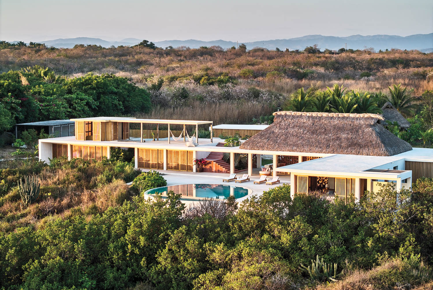 Modern villa with a thatched roof and glass walls nestled in lush greenery, featuring an infinity pool